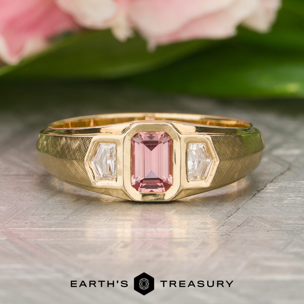 Jeff [custom gallery]: A gold ring featuring a central rectangular pink gemstone and two clear rectangular accent stones, displayed on a textured surface with flowers in the background.
