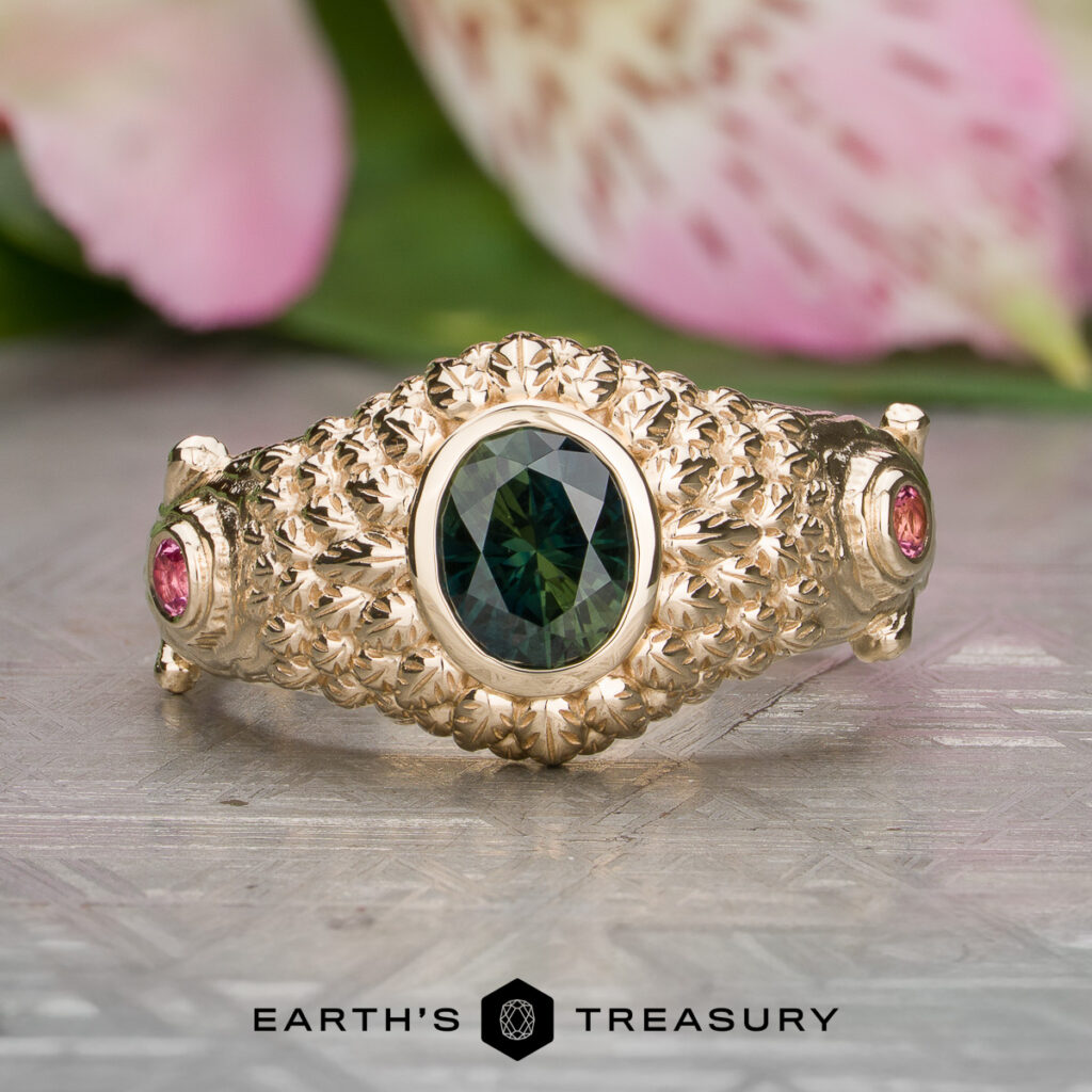 The "Chhabra, Krish [custom gallery]" gold ring showcases an oval green gemstone at the center flanked by two small pink stones, all set in a detailed textured band and shown on a patterned surface.