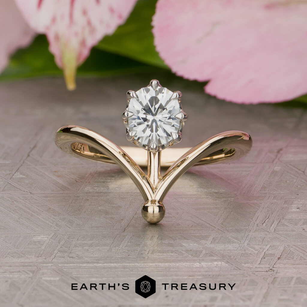 The Paradine Vanessa gold ring features a V-shaped band with a round, faceted gemstone in a prong setting. Pink flower petals and a green leaf are blurred in the background, alongside the Earth's Treasury logo.