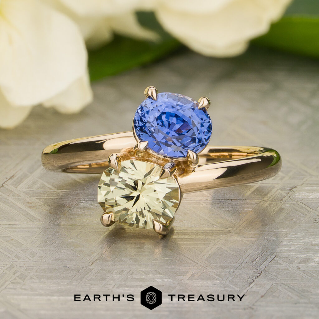 The "Soderberg, Lilly 4 [custom gallery]" gold ring features two round gemstones—one blue and one yellow—set side by side, shown on a textured surface with white flowers in the background.