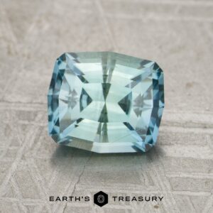 A rectangular, faceted blue-green 2.15-carat Montana Sapphire rests on a textured surface featuring the "Earth's Treasury" logo and text below it.
