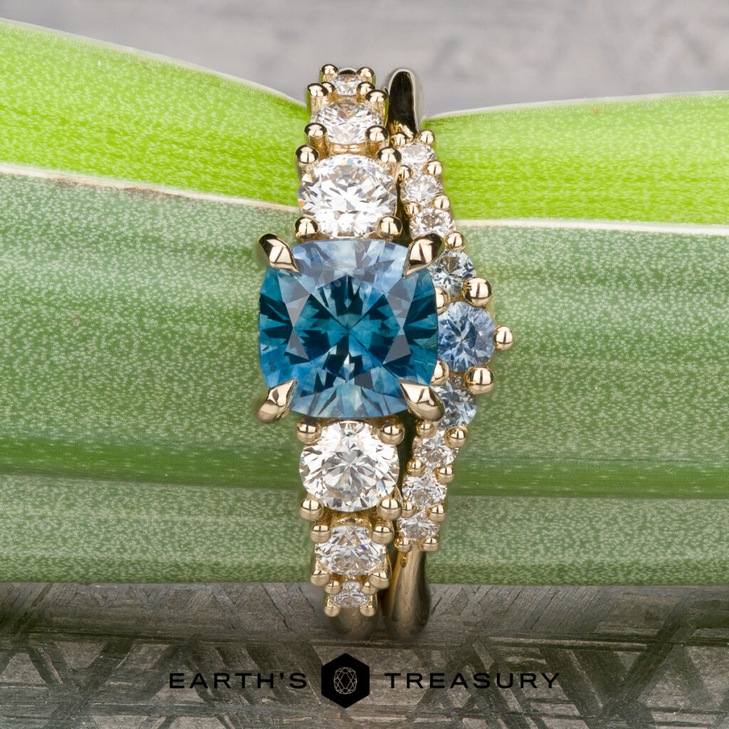 The Buffington, Melissa 1 & 2: a gold ring featuring a square blue gemstone at the center and round white stones on each side, elegantly presented on a green leaf—a stunning women's wedding band choice.