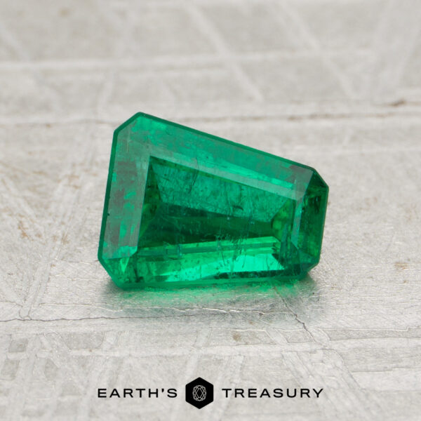 0.78-Carat Zambia Emerald (Oiled) A 0.78-carat Zambia emerald (oiled) with a trapezoid shape is displayed on a textured surface above the Earth's Treasury logo.