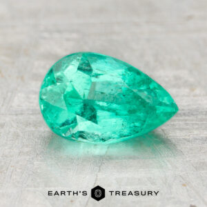 A 1.15-carat pear-shaped, faceted Colombia emerald (oiled) rests on a light textured surface with the text "EARTH'S TREASURY" below.