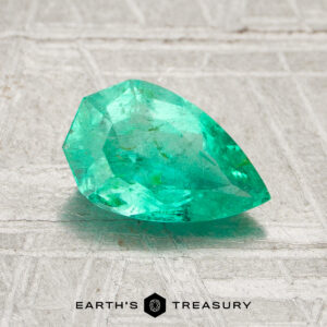 A faceted, pear-shaped 1.08-carat Colombia emerald (oiled) rests on a textured light surface with the "Earth's Treasury" logo and text below.