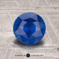A 1.73-carat heated sapphire, round and faceted, stands upright on a textured gray surface with "EARTH'S TREASURY" at the bottom.