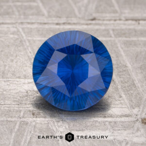 A 1.73-carat heated sapphire, round and faceted, stands upright on a textured gray surface with "EARTH'S TREASURY" at the bottom.