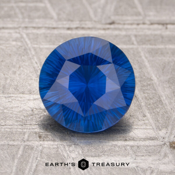 1.73-Carat Sapphire (Heated) A 1.73-carat heated sapphire, round and faceted, stands upright on a textured gray surface with "EARTH'S TREASURY" at the bottom.