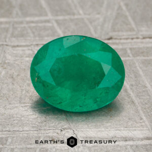 A 1.33-carat oval-cut Colombia emerald (oiled) is displayed on a textured gray surface with "Earth's Treasury" at the bottom.
