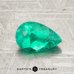 A 1.16-carat Colombia Emerald (oiled), cut in a faceted pear shape, is showcased on a textured gray surface with the "Earth's Treasury" logo at the bottom.