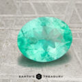 A 1.70-carat Colombia emerald (oiled), cut in a faceted oval shape, rests on a textured gray surface featuring "EARTH'S TREASURY" and a geometric logo below it.