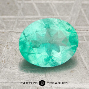 A 1.70-carat Colombia emerald (oiled), cut in a faceted oval shape, rests on a textured gray surface featuring "EARTH'S TREASURY" and a geometric logo below it.
