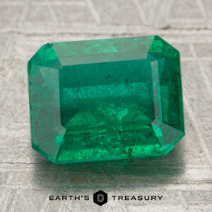 A 2.89-carat Zambia emerald (oiled) in an emerald cut rests on a textured silvery surface, with "Earth's Treasury" shown below.
