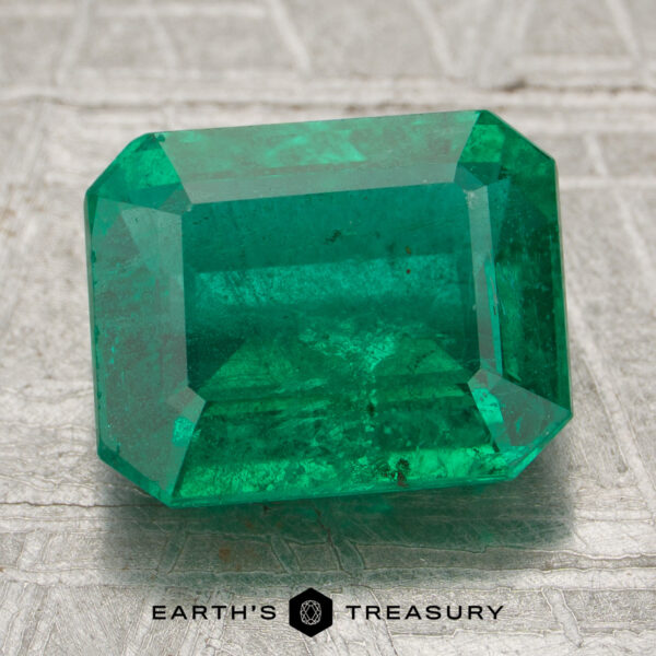 2.89-Carat Zambia Emerald (Oiled) A 2.89-carat Zambia emerald (oiled) in an emerald cut rests on a textured silvery surface, with "Earth's Treasury" shown below.