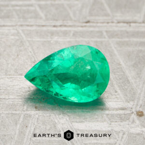 A 0.98-carat pear-shaped green Colombia emerald (oiled) is displayed on a textured light gray surface with the Earth’s Treasury logo at the bottom.