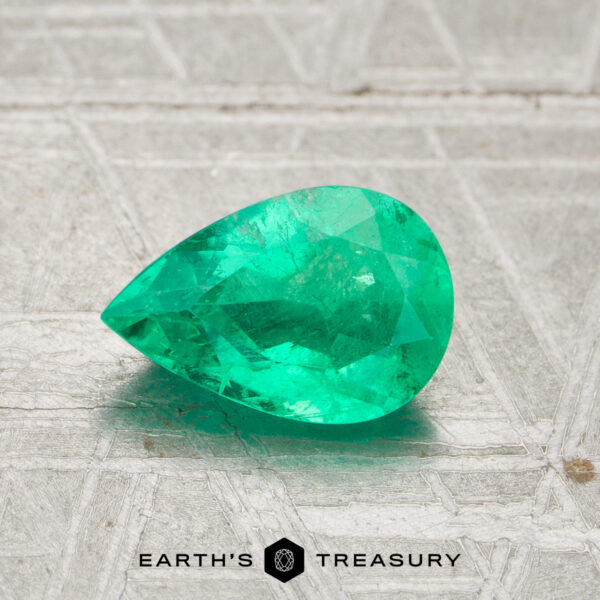 A 0.98-carat pear-shaped green Colombia emerald (oiled) is displayed on a textured light gray surface with the Earth’s Treasury logo at the bottom.