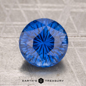 A 2.19-carat heated Ceylon sapphire, faceted and round with a blue hue, rests on a textured gray surface with "Earth's Treasury" below it.