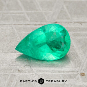 A 0.99-carat pear-shaped Colombia emerald (oiled) rests on a textured gray surface with the "Earth's Treasury" logo beneath it.
