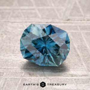 A 1.85-carat heated Montana Sapphire rests on a textured, light-colored surface with "EARTH'S TREASURY" visible below.