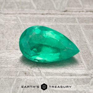 A faceted pear-shaped 0.80-carat Colombia emerald (oiled) rests on a textured surface, with "EARTH'S TREASURY" and a hexagon logo at the bottom.