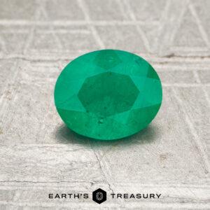 A 0.70-carat Colombia emerald (oiled), faceted and green, rests on a textured metallic surface with "EARTH'S TREASURY" at the bottom.