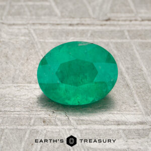 A 0.75-carat Colombia emerald (oiled), oval-cut, is displayed on a textured gray surface with "EARTH'S TREASURY" at the bottom.