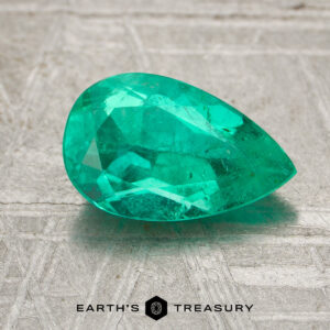 A faceted, pear-shaped 1.24-carat Colombia emerald (oiled) rests on a textured gray surface with the “Earth’s Treasury” logo below.