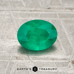 A 0.81-carat oval-cut Colombia emerald (oiled) rests on a textured metallic surface, with "EARTH'S TREASURY" displayed at the bottom.