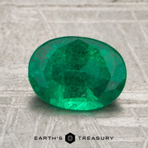A 1.40-carat Zambia emerald (oiled) in an oval cut is displayed on a textured surface, with "Earth's Treasury" and a hexagon logo beneath it.