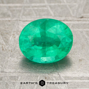 A 1.54-carat oval-cut Zambia emerald (oiled) is displayed on a textured gray surface with the "Earth's Treasury" logo at the bottom.
