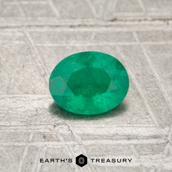 A 0.44-carat Colombia Emerald (oiled), oval-shaped and green, is displayed on a textured silvery surface with "EARTH'S TREASURY" at the bottom.