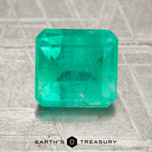 A 1.65-carat oiled Colombia emerald with a rectangular cut is displayed on a textured surface, featuring the "Earth's Treasury" logo and text at the bottom of the image.