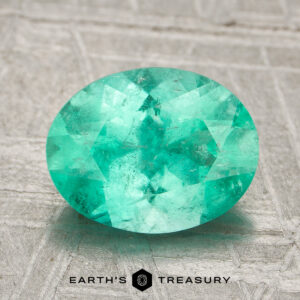 A 1.89-carat, oval-cut Colombia emerald (oiled) rests on a textured surface, with "Earth's Treasury" and a geometric logo displayed beneath it.