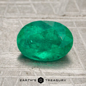 A 1.36-carat Colombia emerald (oiled), oval-cut, is displayed on a textured metallic surface with the "Earth's Treasury" logo beneath.