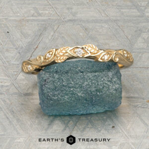 The "Amborella" band in 14K yellow gold with leaf and diamond accents is shown on a rough blue gemstone, with the "Earth's Treasury" logo and text below.