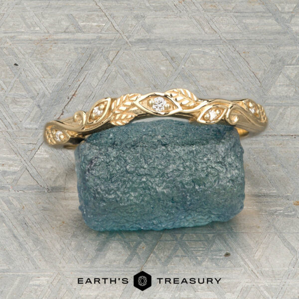 The "Amborella" band in 14K yellow gold The "Amborella" band in 14K yellow gold with leaf and diamond accents is shown on a rough blue gemstone, with the "Earth's Treasury" logo and text below.