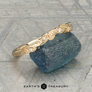 The "Amborella" band in 14K yellow gold with leaf and diamond accents is shown on a rough blue gemstone, with the "Earth's Treasury" logo and text below.