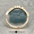 The "Amborella" band in 14K yellow gold with leaf and diamond accents is shown on a rough blue gemstone, with the "Earth's Treasury" logo and text below.