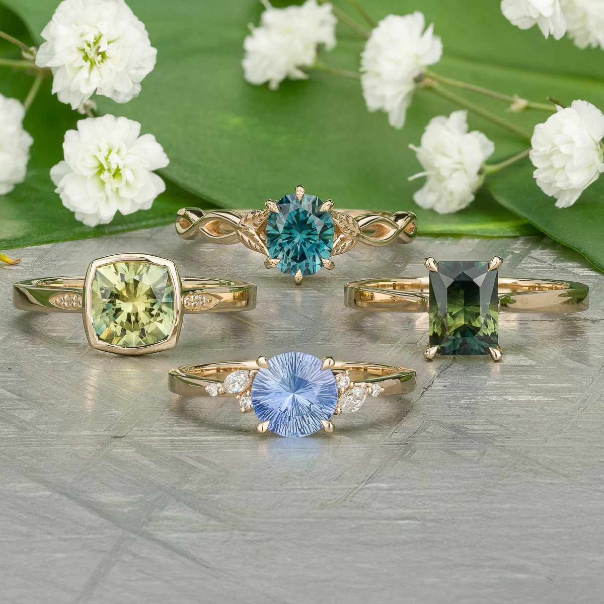 Four "Stacked from 76 images. Method=B (R=14,S=2)" gold rings with green, blue, and teal gemstones are displayed on a silver surface with white flowers in the background.