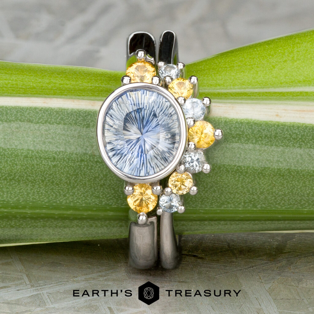 The Rheault, Nicole (Nikki) 1 is a silver ring with an oval blue gemstone beside yellow and white stones, shown on a green leaf background with the Earth’s Treasury logo—ideal for those wanting unique women’s wedding bands.