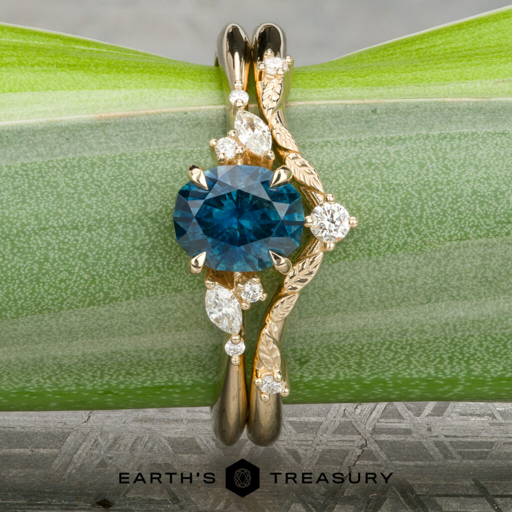 The "Dean, Brian" ring features a blue oval gemstone with diamond accents on a gold band, shown atop a green leaf. Ideal for women's wedding bands, the Earth's Treasury logo appears at the bottom.
