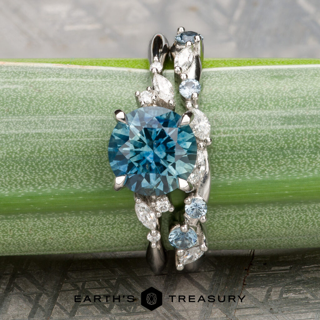 The "Yamada, Iris and Lok Wan, Kai 1 & 2" features a round blue gemstone ring with a matching curved band in white metal and accent stones, displayed on a green stem. Ideal for women’s wedding bands. Earth's Treasury logo shown at bottom.