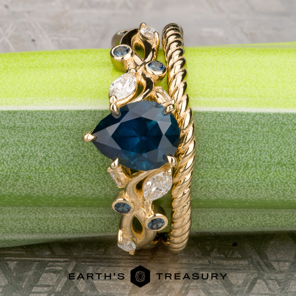 The Mathau, Andrew 1 & 2 set features a gold ring with a blue pear-shaped gemstone, diamond accents, and a twisted gold band. Displayed on green, it’s ideal for those seeking unique women's wedding bands. Earth's Treasury logo appears below.