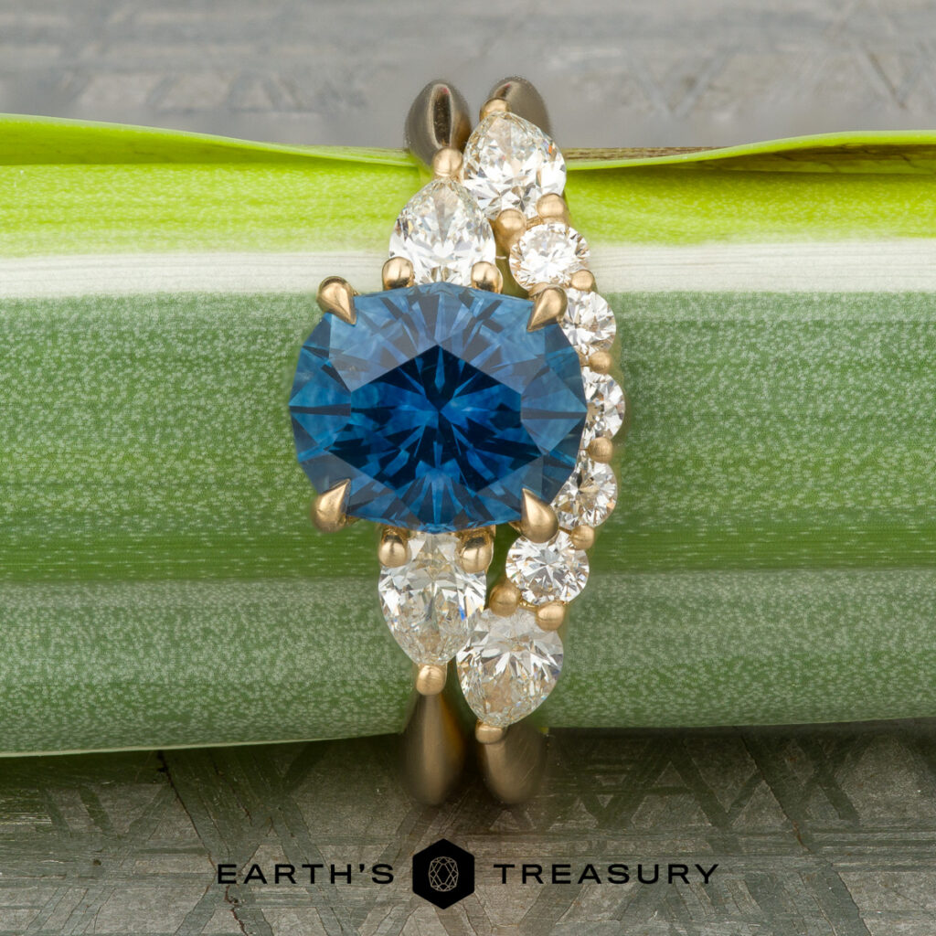 A gold ring by Miller, Ryan features an oval blue gemstone with five clear round and pear-shaped stones beside it, displayed on a green leaf. The “Earth’s Treasury” logo appears at the bottom, adding a special detail to this women’s wedding band.