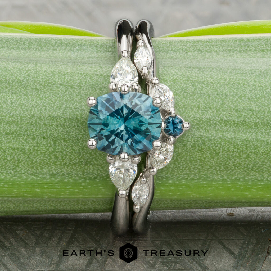 A platinum women's wedding band by Kofahl, Dustin features a round blue gemstone, a smaller blue stone, and four marquise-cut white gems. The ring is displayed on a green surface with the "Earth's Treasury" logo below.
