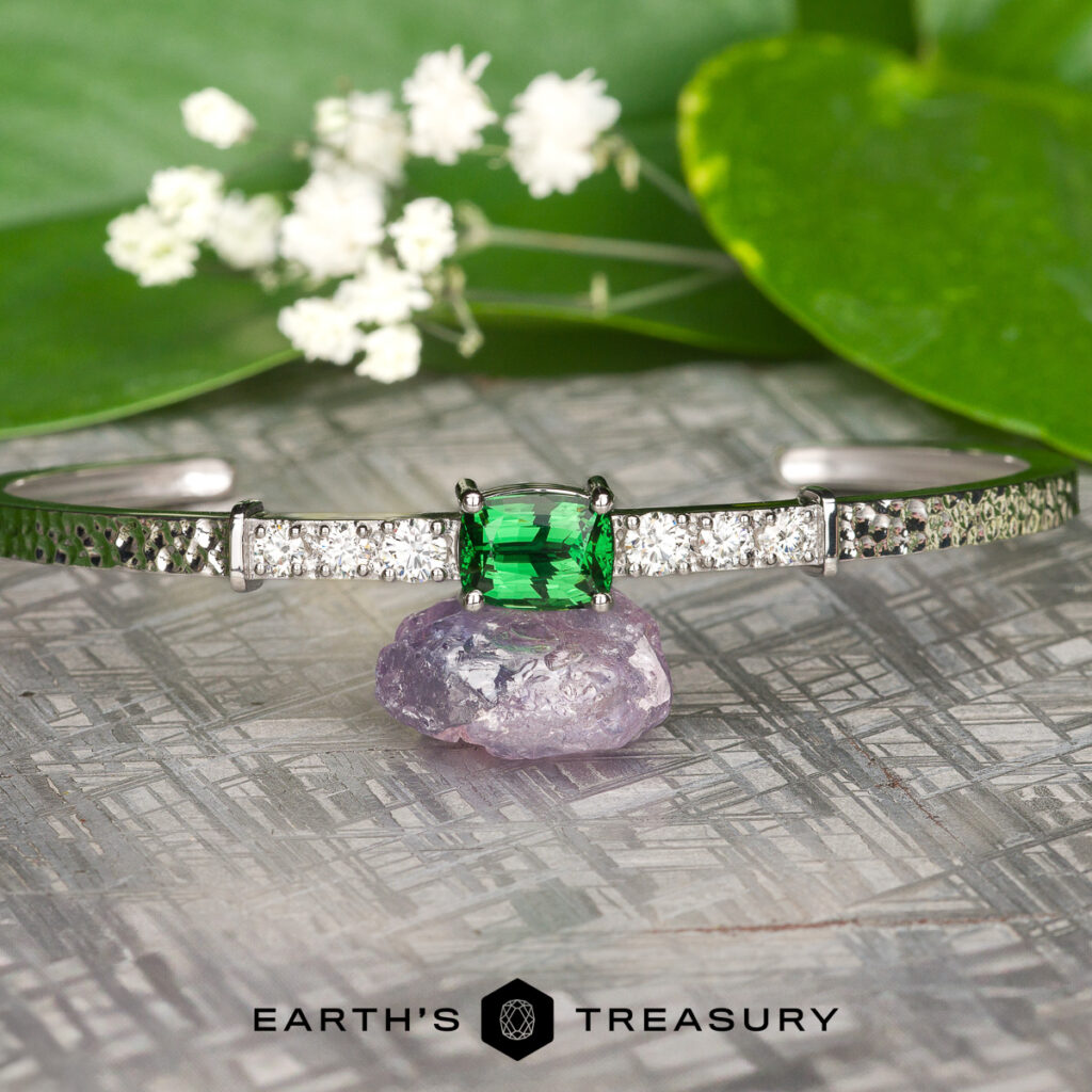 The "shone, john 28 [custom gallery]" silver bracelet with a green gemstone centerpiece and side diamonds is displayed on a purple crystal, accented by green leaves and white flowers. "Earth’s Treasury" logo appears at the bottom.