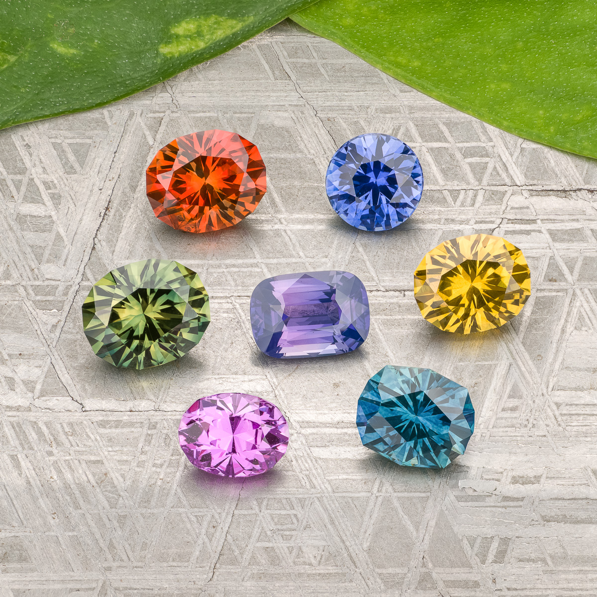 Seven faceted gemstones in red, blue, yellow, green, pink, purple, and teal are displayed on a patterned gray surface with two green leaves above. Product: "Stacked from 45 images. Method=B (R=7,S=2).