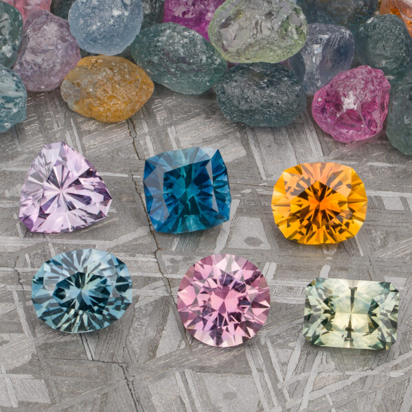 Six faceted gemstones in various colors, including stunning Montana Sapphires, are shown on a textured surface with uncut stones behind them. Product: Stacked from 58 images. Method=B (R=7,S=2).