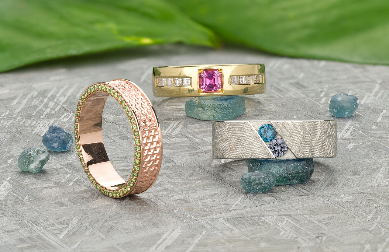 Shop-Gemstone-Bands-scaled-1 features three rings on a textured surface: a rose gold with green gems, a yellow gold with diamonds and a pink center stone, and a silver band adorned with blue and white gems.
