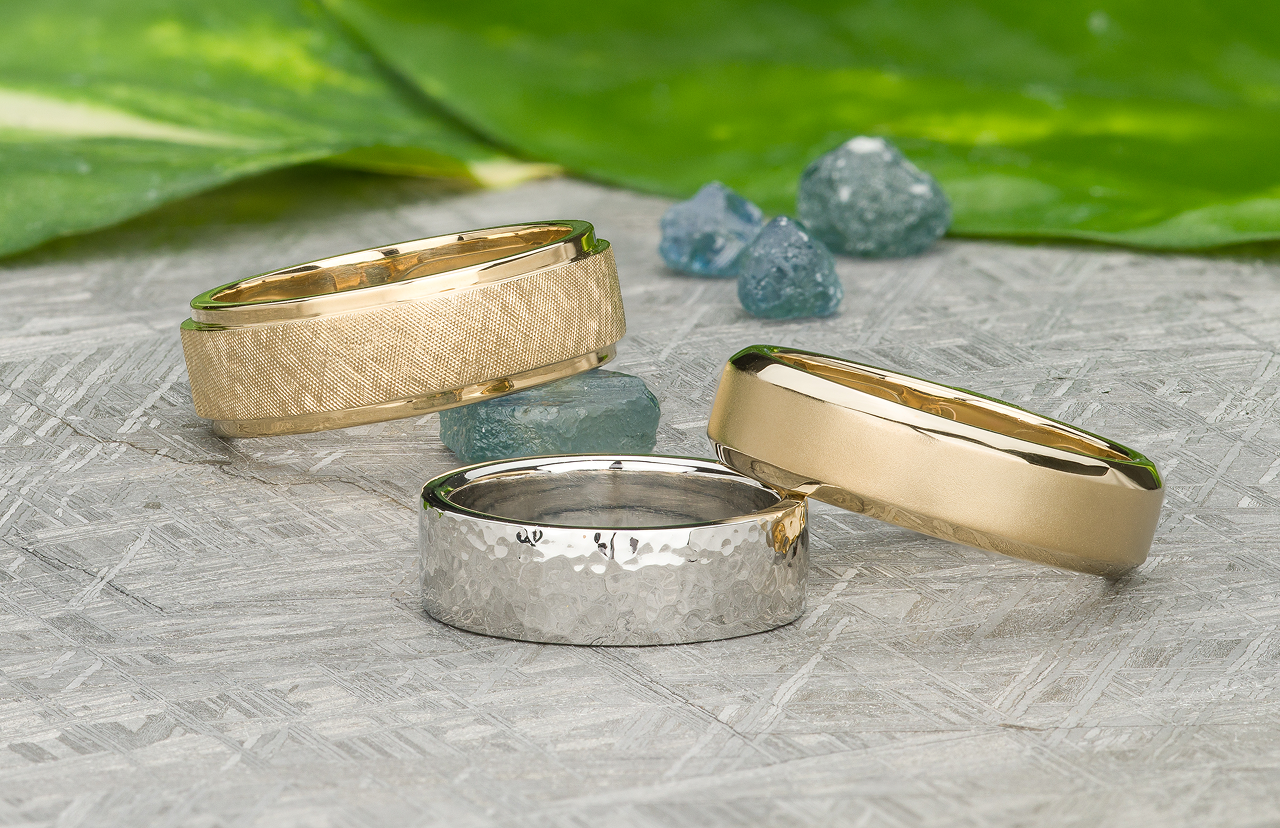 Three Shop-Plain-Bands-scaled-1 wedding bands—two gold and one silver—are displayed on a textured surface, surrounded by blue stones and green leaves in the background.